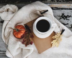 Close-up of a miniature pumpkin and fake leaves on a white blanket, on a rustic wood table. There's a coffee mug with coffee in it sitting on top of a brown notebook, on the blanket.