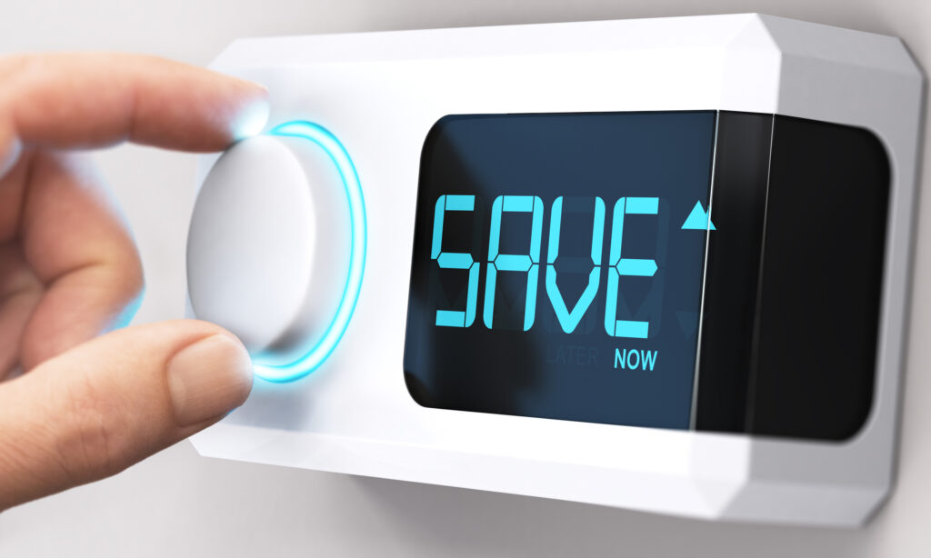 Hand turning a thermostat knob to increase savings by decreasing energy consumption. Digital display shows "SAVE now" in blue letters.